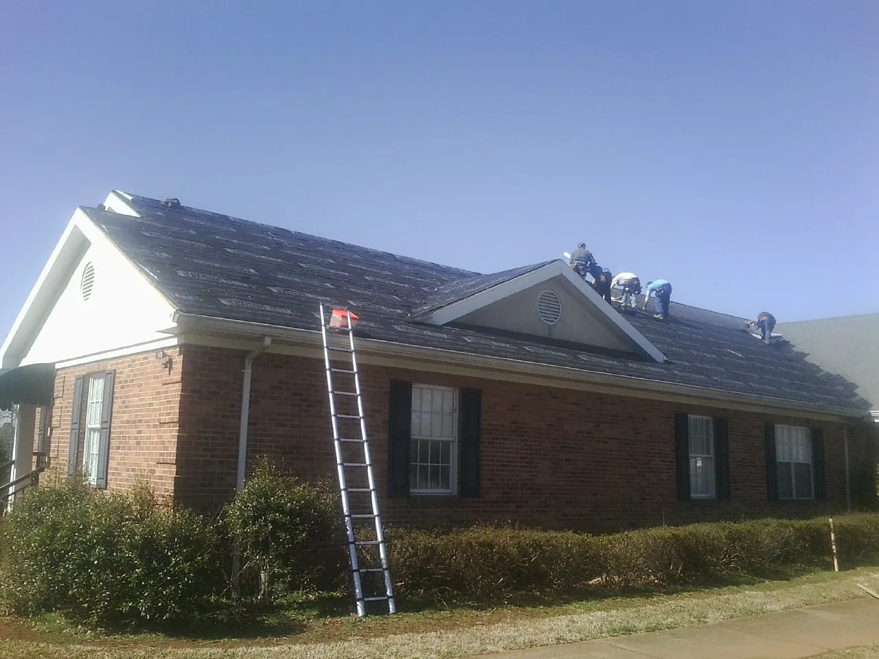 Crew tearing shingles off a brick ranch roof with a ladder set — Emergency Roof Repair work by our crew in Dolomite