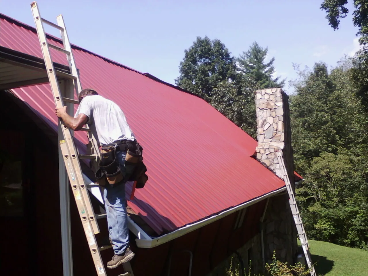 Roofer on a ladder beside a red metal roof and stone chimney — Soffit & Fascia Repair by Roofing Brothers in Dolomite